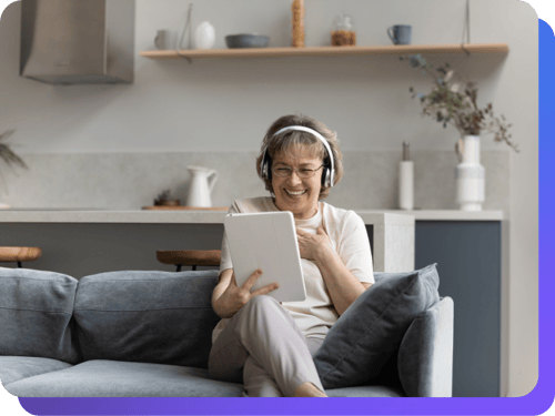 Smiling older woman wearing headphones, sitting on a couch and holding a tablet during a video call, representing the joy of sending and receiving digital greeting cards. (1)