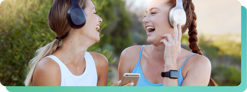 Two women in athletic wear smiling and laughing outdoors while listening to music on wireless headphones and holding a smartphone (1)