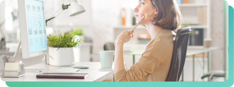 Woman in a beige cardigan sitting at a desk, holding a pen to her chin while reviewing mobile app wireframes on a desktop monitor
